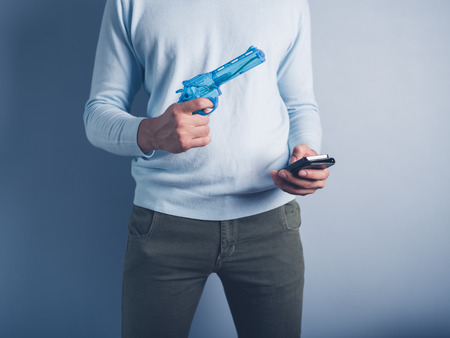 A young man is standing against a blue background with a water pistol and a smart phoneの写真素材