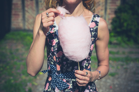 A young woman wearing a dress is standing outside an old farmhouse with a candyfloss on a sunny summer dayの写真素材