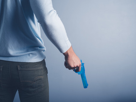 A young man is standing against a blue background and is posing with a water pistolの写真素材