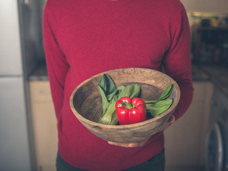 A young man is standing in a kitchen with a bowl of pak choi and red pepperの写真素材