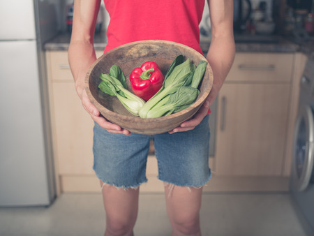 A young woman is standing in a kitchen with a bowl of pak choi and red pepperの写真素材