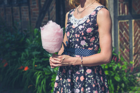 A young woman wearing a dress is standing outside an old farmhouse with a candyfloss on a sunny...