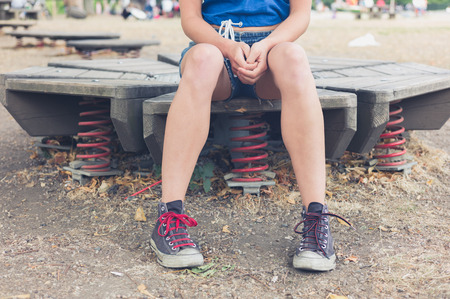 A young woman is sitting on some playground equipment with springs outside in summerの写真素材