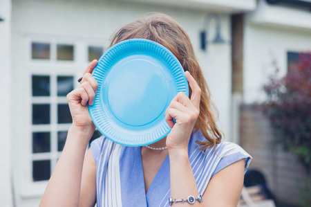 A young woman is hiding her face behind a blue paper plate in a gardenの写真素材
