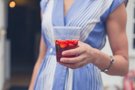 A young woman wearing a summer dress is standing in a garden with a drink in her hand