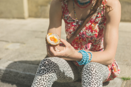 A young woman is sitting on the curb on the street and is eating a scotch eggの写真素材