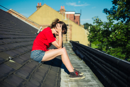 A sad young woman is sitting on a roof with her head in her handsの写真素材
