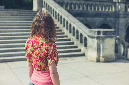 A young woman is standing near the steps of an old Victorian building outside in a park or grand formal gardenの写真素材
