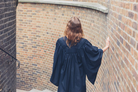 A young woman wearing a graduation gown is walking down some stairs outside by a brick wall