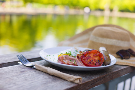 A cooked breakfast with sausage bacon and eggs on a plate next to a hat by a pond in the parkの写真素材