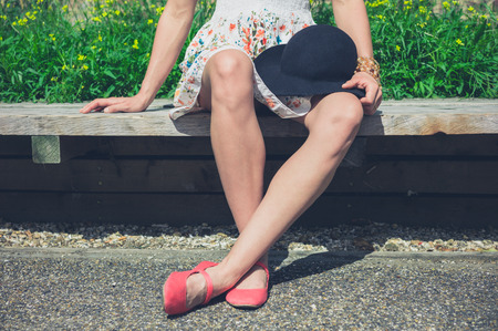 A young woman wearing a summer dress is sitting on a bench outside with a hatの写真素材