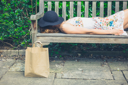 A young woman wearing a summer dress and a hat is relaxing on a bench in the park on a sunny day, there is a paper bag next to herの写真素材