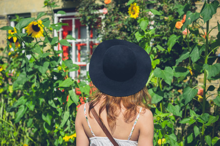 A young woman wearing a hat and a dress is admiring some sunflowers outside a house on a sunny day in summerの写真素材