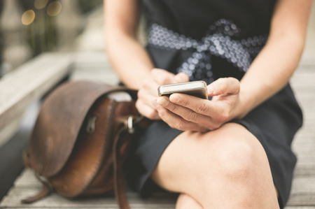 A stylish young woman is sitting on a bench in the city and is using her smart phoneの写真素材