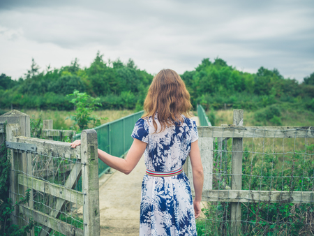 A young woman wearing a dress is opening a gate in the countryside on a gloomy and cloudy dayの写真素材