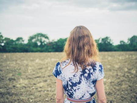 A young woman wearing a dress is standing in a barren field on a cloudy and gloomy dayの写真素材