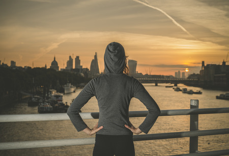 A young woman wearing a sporty hoodie is standing on a bridge in London at sunriseの写真素材