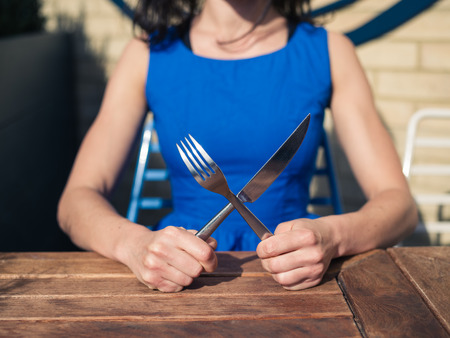 A young woman wearing a blue dress is sitting at a table outside a restaurant with a knife and fork in her handsの写真素材
