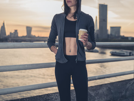 An athletic young woman with toned abs  is standing on a bridge in London at sunrise with a paper...