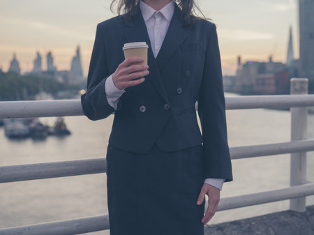 A young businesswoman is standing on a bridge with a cup of coffee in her hand and is admiring the sun rise over the London skylineの写真素材