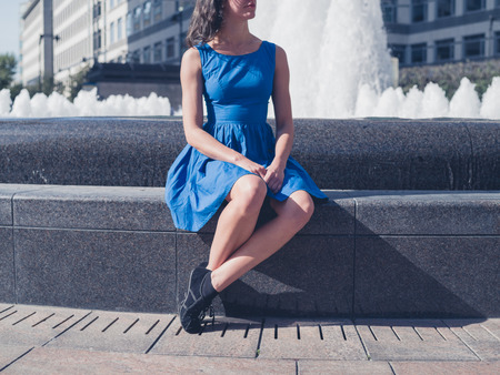 A young woman wearing a blue dress is sitting by a fountain in the city on a sunny dayの写真素材