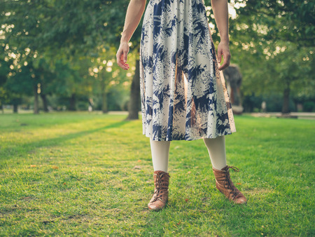 A young woman wearing a dress is standing in a park on the grass at sunsetの写真素材