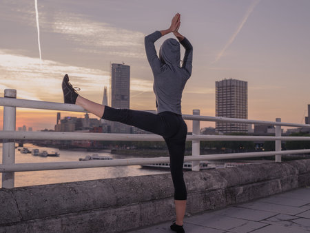 A young fitness woman is standing in a yoga pose on a bridge in London at sunriseの写真素材
