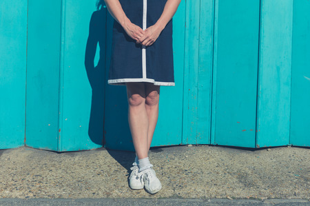 A beautiful young woman wearing a dress is standing by a big blue garage door on a sunny dayの写真素材