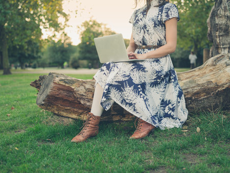 A young woman is sitting on a log in the park at sunset and is working on her laptop computerの写真素材