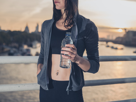 A fit and athletic young woman is standing with a bottle of water on a bridge in London at sunriseの写真素材