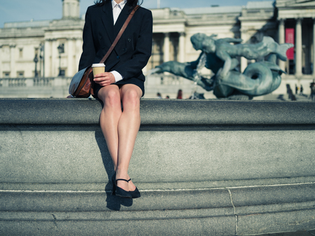 A young businesswoman is sitting by a fountain in a city and is drinking coffeeの写真素材