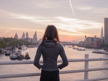 A young woman wearing a sporty hoodie is standing on a bridge in London at sunriseの写真素材