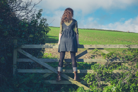 A young woman is standing on a gate in the countryside and is looking at the landscape on a sunny dayの写真素材