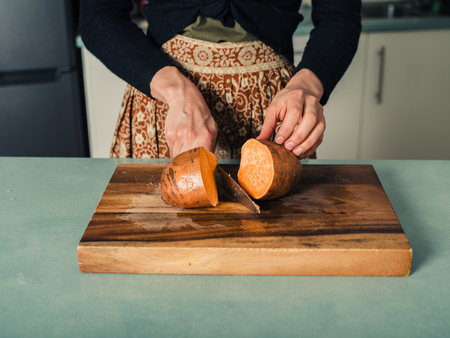 A young woman is in a kitchen and is cutting a sweet potato in halfの写真素材