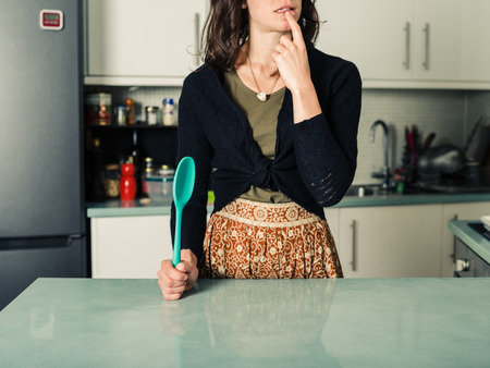 A young woman is standing in a kitchen with a spoon and is looking confusedの写真素材