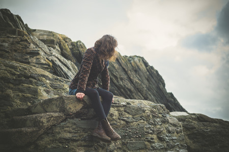 A young woman is sitting on a rock on the coast and is admiring the nature on an overcast dayの写真素材