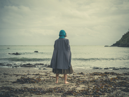 A young barefoot woman wearing a headscarf is standing on a beach on a cold and windy dayの写真素材