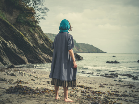 A young barefoot woman wearing a headscarf is standing on a beach on a cold and windy dayの写真素材