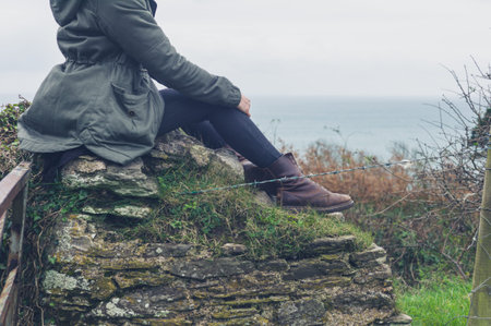 A young woman wearing a winter coat is resting on a wall in the countryside