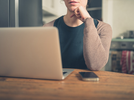 A young woman is sitting in her kitchen and is using her latop and a smart phoneの写真素材