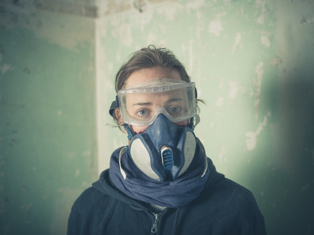 A young woman is wearing a dust mask and protective goggles is standing in a derelict room undergoing renovationsの写真素材