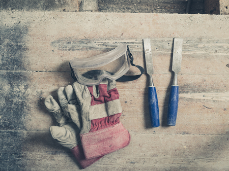 A pair of workman gloves two chisels and a pair of protective goggles on a wooden floorの写真素材