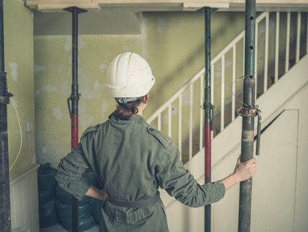 A young woman wearing a hard hat and a boiler suit is inspecting some acro props supporting the...