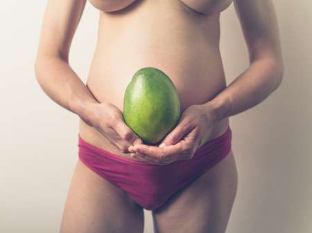 A young pregnant woman is holding a mango to demonstrate the current size of her unborn babyの写真素材