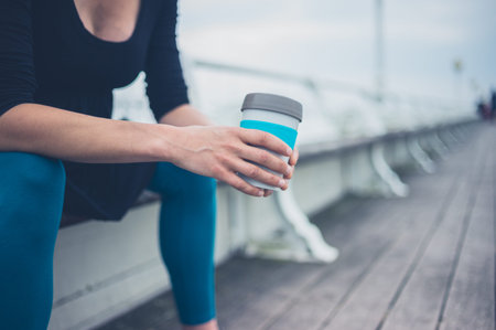 A young woman with a cup is sitting on a bench on the pier