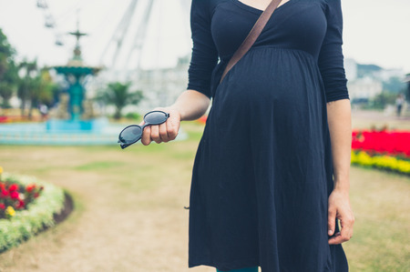 A pregnant woman is standing in a park with her sunglasses in her handの写真素材