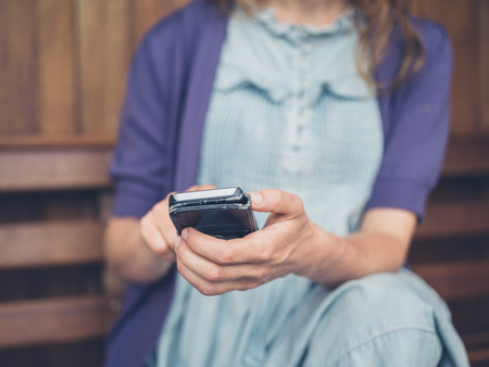 A young woman is sitting on a bench outside and is using a smartphoneの写真素材
