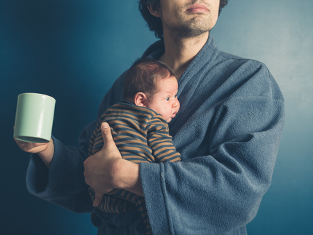 A young father in a bathrobe is holding a mug and his baby sonの写真素材