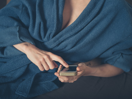 A woman wearing a bathrobe is lying in bed and is using a smartphoneの写真素材