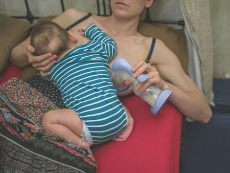 A young mother in bed is breastfeding her baby whilst expressing milk with a pump